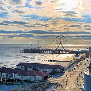 May include: A coastal scene at sunset with a Ferris wheel, pier, and buildings along a promenade. The sky is filled with clouds, and the sun shines brightly over the sea. People are walking along the boardwalk.
