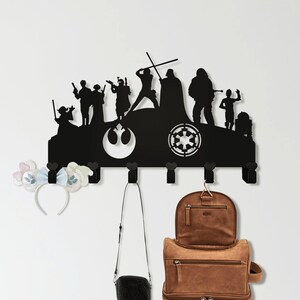 May include: Black metal wall-mounted coat rack featuring a Star Wars themed silhouette of characters and symbols. The rack has multiple hooks holding a small black purse, a brown leather backpack, and a smaller matching bag. A floral headband hangs on the left.
