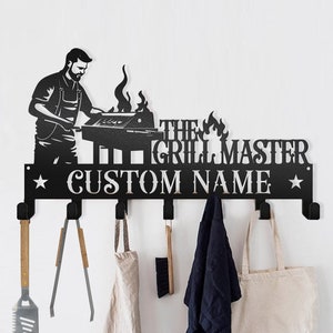 May include: Black metal wall-mounted coat rack with a grill master design. The rack has six hooks and features the text "The Grill Master" and "Custom Name".