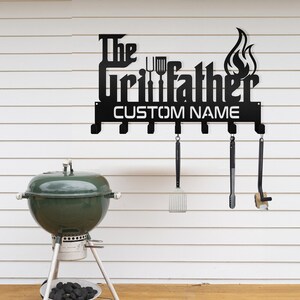 May include: Black metal wall-mounted grill tool rack with the text "The Grillfather" and a customizable name space. The rack has six hooks for hanging grilling tools. A green grill is in the background.