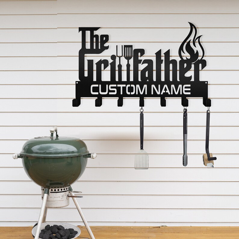 May include: Black metal wall-mounted grill tool rack with the text "The Grillfather" and a customizable name space. The rack has six hooks for hanging grilling tools. A green grill is in the background.