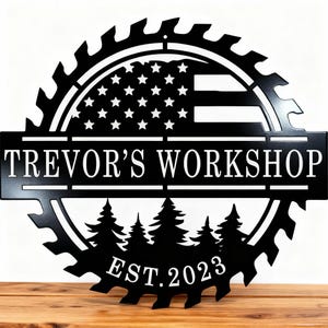 Può includere: Un'insegna da parete circolare in metallo nero a forma di lama di sega. La metà superiore presenta il design di una bandiera americana, mentre la metà inferiore mostra una foresta di pini. Il testo recita TREVOR'S WORKSHOP e EST. 2023.
