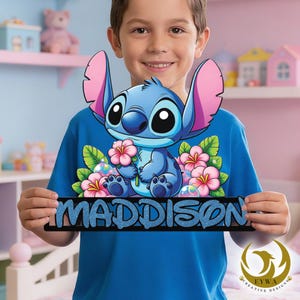 May include: A blue t-shirt with a cartoon character holding flowers, and the name "MADDISON" in glittery blue letters on a black sign. The character has large ears and is surrounded by pink flowers. The shirt is worn by a person.