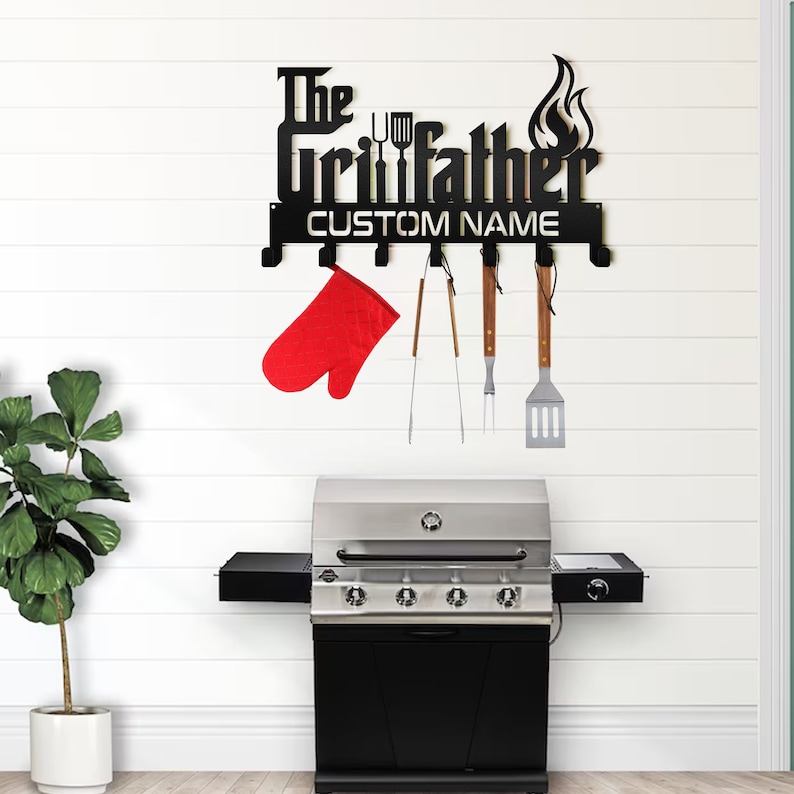 May include: Black metal grill tool rack with the text 'The Grillfather' and a flame design. The rack has hooks for hanging grilling tools. A red oven mitt and grilling tools are hanging from the hooks. A black and silver gas grill is in the background.