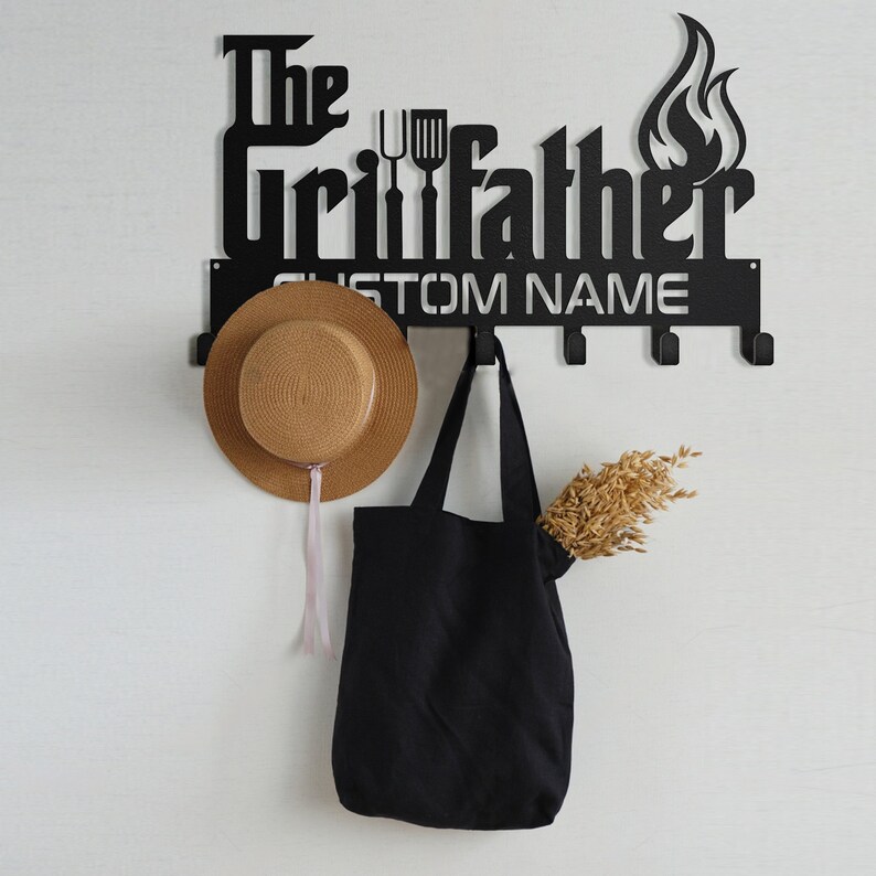 May include: Black metal wall-mounted coat rack with the text "The Grillfather" and a flame design. The rack has five hooks and a space for a custom name. A straw hat, a black tote bag, and a bundle of dried wheat are hanging on the hooks.
