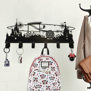 Puede incluir: Perchero de pared de metal negro con forma de horizonte de parque temático de Disney con 7 ganchos. Una mochila de Mickey Mouse blanca y roja cuelga de un gancho, y una bolsa de cuero marrón cuelga de otro gancho.