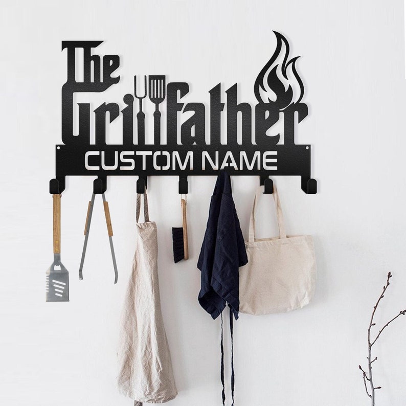 May include: Black metal wall mounted coat rack with the words 'The Grillfather' and 'CUSTOM NAME' in black lettering. The rack has six hooks and is decorated with a flame design. The rack has a spatula, tongs, an apron, a brush, a tote bag and a jacket hanging on the hooks.