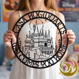 Puede incluir: Arte de pared circular de metal negro con una silueta detallada del Castillo de Hogwarts, rodeada por las palabras "CASA HOGWARTS WELCOMES YOU HOME". El diseño incluye árboles, estrellas y una luna. La obra es sostenida por una persona.
