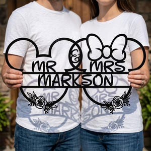 May include: Black metal Mr. and Mrs. sign with the last name "Markson" in the center. The design incorporates Mickey Mouse ears and a bow, with floral accents. A unique wedding or anniversary decoration.