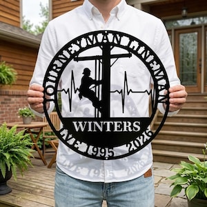 May include: A black metal sign with the words "JOURNEYMAN LINEMAN" in a circular design. The sign features a silhouette of a lineman on a utility pole, with the name "WINTERS" and the years "1995" and "2026".
