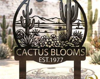 May include: Black metal garden sign featuring a desert landscape with cacti, flowers, and mountains. The sign reads "CACTUS BLOOMS EST.1977" and is designed with a circular frame and two stakes for ground placement. A decorative piece for outdoor spaces.