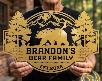 May include: A gold-colored metal sign featuring a bear, mountains, and trees. The sign reads "BRANDON'S BEAR FAMILY" and "EST. 2026". The sign is held by a person.