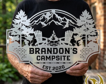 May include: A silver metal sign featuring a laser-cut design of two chairs, a fire pit, pine trees, and mountains. The text reads BRANDON'S CAMPSITE and EST. 2026. The sign has a decorative border with a brick pattern and barbed wire detail.