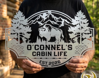 May include: A silver-colored metal wall sign featuring a laser-cut design of a bison standing in front of mountains and pine trees. The text O'CONNEL'S CABIN LIFE and EST. 2026 is cut into the metal, framed by a decorative border with brick patterns.
