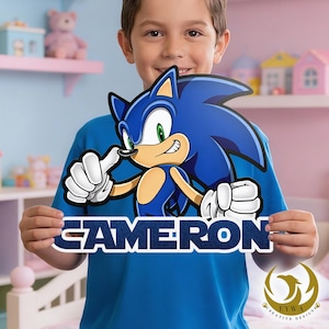 May include: A blue sign featuring the Sonic the Hedgehog character with the name "CAMERON" in white letters. The sign is held by a person wearing a blue shirt. The Sonic character is blue, tan, and white, with a thumbs-up gesture.