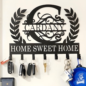 May include: Black metal key holder with the text "HOME SWEET HOME" and the name "CARDANY". The key holder has a large letter "C" at the top, surrounded by a laurel wreath design, and several hooks for keys.