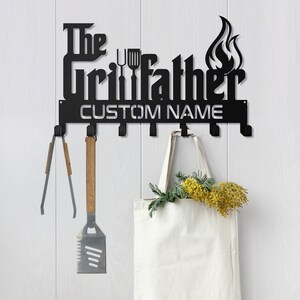 May include: Black metal wall-mounted hook rack with the text "The Grillfather" and a flame design. The rack has six hooks and a space for a custom name. A spatula, tongs, and a tote bag are hanging on the hooks.