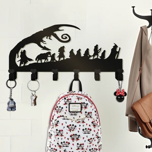 May include: Black metal wall-mounted coat rack with a silhouette of characters from the Lord of the Rings movie. The rack has six hooks and is decorated with a tree and a dragon. A backpack, keys, and a small stuffed animal are hanging on the hooks.