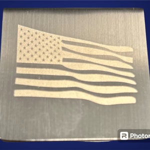 May include: A silver metal rectangle with an etched image of the American flag. The flag is white and has a wavy appearance.