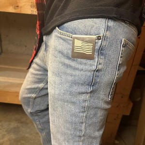 Op de afbeelding: Een persoon draagt ​​een blauwe jeans met een kleine metalen Amerikaanse vlag sticker in de voorzak.