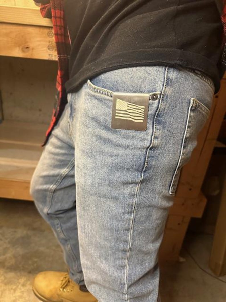 Op de afbeelding: Een persoon draagt ​​een blauwe jeans met een kleine metalen Amerikaanse vlag sticker in de voorzak.