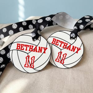 Puede incluir: Dos etiquetas blancas con forma de voleibol con el nombre "BETHANY" y el número "11" en rojo. Las etiquetas están unidas a una cinta negra con voleiboles blancos. El fondo es de tela beige claro.
