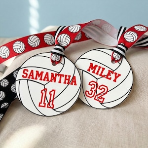 Pode incluir: Dois ornamentos circulares brancos em forma de bolas de voleibol com linhas pretas. Um apresenta o nome SAMANTHA e o número 11 a vermelho, enquanto o outro exibe MILEY e 32 a vermelho. Ambos pendurados por uma fita vermelha com padrões de voleibol.