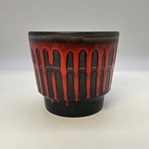 May include: A ceramic planter with a matte black base and a textured surface featuring a vibrant red glaze that drips vertically from the rim. The vessel has a wide, open top that tapers down to a narrower, solid black circular base.