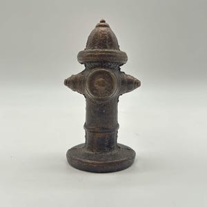 May include: A small, dark brown, textured cast iron fire hydrant figurine. The object features a rounded top, a central circular nozzle, and two side outlets, all set upon a wide, circular base. The surface has a rough, weathered appearance.