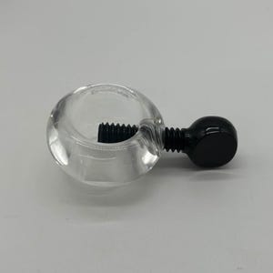 May include: A clear glass nipple clamp with a black screw and a black ball end. The clamp is designed to be adjustable. The product is shown on a white background.