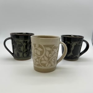 May include: Three ceramic mugs with handles. The mugs are in shades of black and beige, with floral patterns. The central mug is beige with a white floral design. The other two mugs are black with a darker floral pattern.
