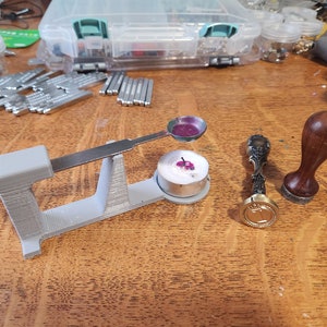 Può includere: Una stazione di fusione per sigilli di cera stampata in 3D con un cucchiaio di metallo, una candela e un timbro per sigilli di cera in legno e metallo.