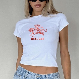 Könnte beinhalten: Weißes, bauchfreies T-Shirt mit einem roten Motiv einer Frau, die auf einem Tiger reitet. Der Text "HELL CAT" ist unter dem Motiv gedruckt.