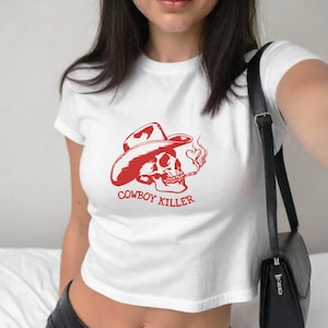 May include: White cropped t-shirt with a red graphic of a skull wearing a cowboy hat and smoking a cigarette. The text "COWBOY KILLER" is printed below the skull.