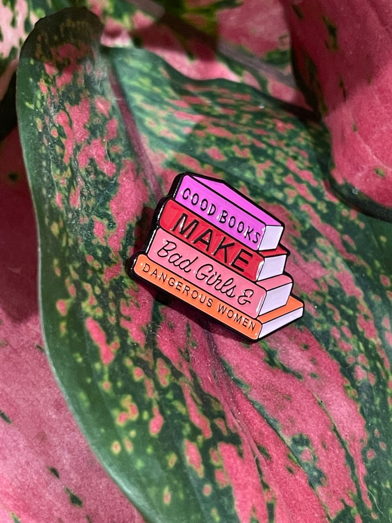 May include: A colorful enamel pin shaped like a stack of books. The top book is pink and reads "GOOD BOOKS". The next book is red and says "MAKE". The third book is pink and says "BAD GIRLS &". The bottom book is orange and says "DANGEROUS WOMEN".