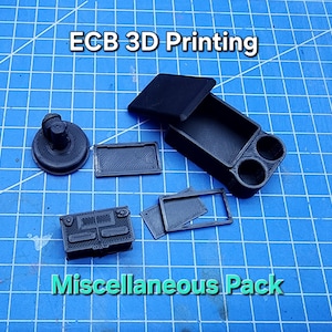 以下が含まれることがあります： 雑多なパック用の3Dプリントされた黒いプラスチック部品のセット。部品には、蓋付きの小さな箱、小さな長方形の部品、小さな正方形の部品、ノブ付きの丸い部品が含まれています。