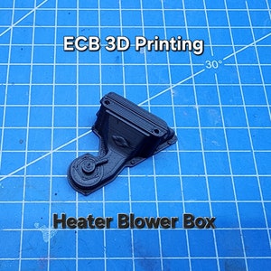 以下が含まれることがあります： 黒色の3Dプリントされたヒーターブロワーボックス。ボックスにはファンと長方形のハウジングがあります。ボックスの上に「ECB 3D Printing」というテキストが表示されています。