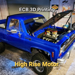 Peut inclure: Un camion miniature bleu avec le capot ouvert, révélant un moteur détaillé. Le camion a un arceau de sécurité noir et un autocollant noir et blanc sur le pare-brise qui dit "RC 4x4". Le camion est posé sur une surface grise avec un tapis vert et noir en arrière-plan. Le texte "ECB 3D Printing Garage" est visible sur une pancarte au-dessus du camion. Le texte "High Rise Motor" est visible sous le camion.
