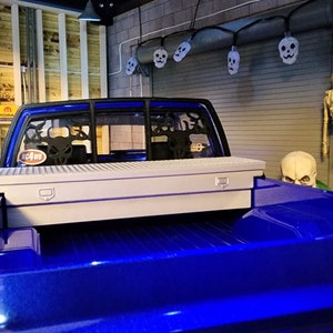 Puede incluir: Una camioneta azul con una caja de herramientas blanca en la caja. La camioneta tiene una jaula antivuelco negra con un diseño de calavera. La camioneta está estacionada en un garaje con una pared de madera y una pared de ladrillo.