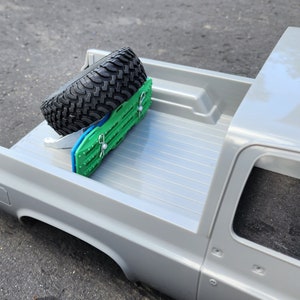 Puede incluir: Una caja de camioneta plateada con una rueda de repuesto asegurada con una correa de rueda verde y azul.