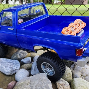Puede incluir: Una camioneta modelo azul brillante con neumáticos todoterreno grandes, estacionada sobre una superficie rocosa. La caja de la camioneta contiene una tabla de tracción naranja. La camioneta tiene un diseño detallado, que incluye detalles cromados y un portón trasero rebajado.