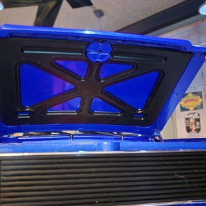 Puede incluir: Un camión de juguete azul con el capó abierto, que revela un compartimento del motor negro con una parrilla de plástico azul. El camión tiene una parrilla negra y faros plateados.