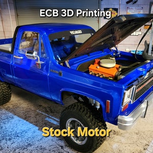 Może przedstawiać: Niebieski pick-up Chevrolet z lat 70. z otwartą maską, ukazującą silnik. Samochód ma czarne wnętrze i czarną kratkę. Samochód stoi na betonowej podłodze w garażu. Tekst "ECB 3D Printing" jest widoczny na znaku w tle. Tekst "Stock Motor" jest widoczny na pierwszym planie.