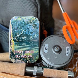 May include: A close-up of a fly fishing tackle box with a colorful image of a river and the text "Piney River Fly Box Co.". The box is sitting on a wooden surface next to a fly fishing reel and a fishing rod.