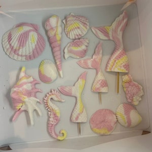 Könnte beinhalten: Ein Set mit 13 rosa und gelben Meerjungfrauen-Schwanz- und Muschel-Kuchen-Toppern. Die Topper sind aus Fondant und haben ein glitzerndes Finish.