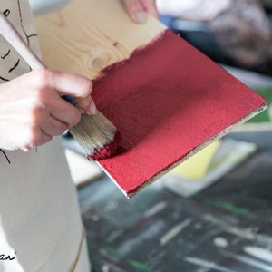 Puede incluir: Una persona aplica pintura roja a una tabla de madera con un pincel. La tabla est&aacute; parcialmente pintada, revelando la veta natural de la madera. La imagen presenta el logotipo de Annie Sloan en la esquina inferior izquierda.
