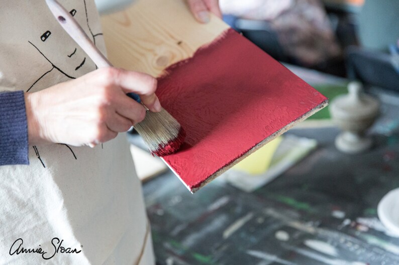 Puede incluir: Una persona aplica pintura roja a una tabla de madera con un pincel. La tabla est&aacute; parcialmente pintada, revelando la veta natural de la madera. La imagen presenta el logotipo de Annie Sloan en la esquina inferior izquierda.