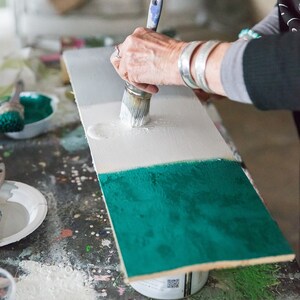 Puede incluir: Una persona pinta una tabla de madera con un pincel. La tabla est&aacute; dividida en tres secciones, pintadas de blanco, gris y turquesa. El logotipo de Annie Sloan es visible en la esquina inferior izquierda.