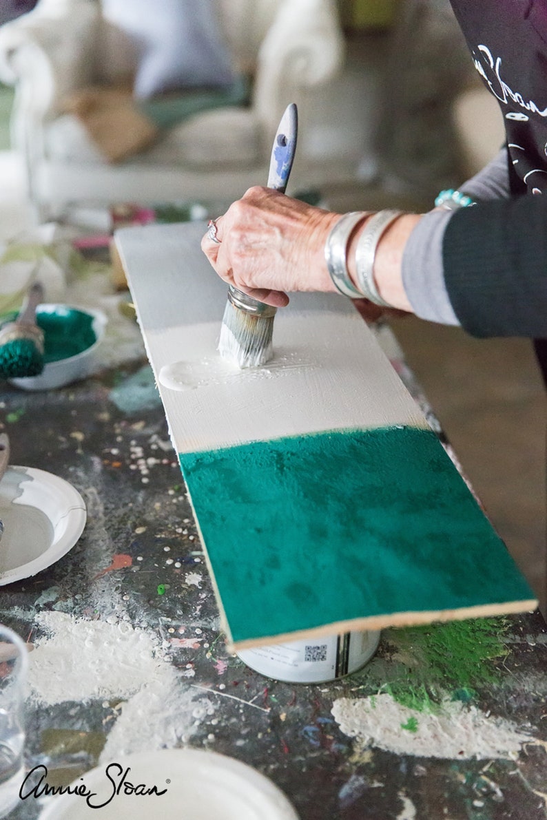 Puede incluir: Una persona pinta una tabla de madera con un pincel. La tabla est&aacute; dividida en tres secciones, pintadas de blanco, gris y turquesa. El logotipo de Annie Sloan es visible en la esquina inferior izquierda.