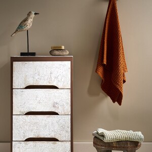 May include: A white dresser with four drawers and brown trim sits on a white floor. A small wooden stool with a woven cushion sits in front of the dresser. A brown towel hangs on a hook on the wall. A small wooden bird figurine sits on top of the dresser.
